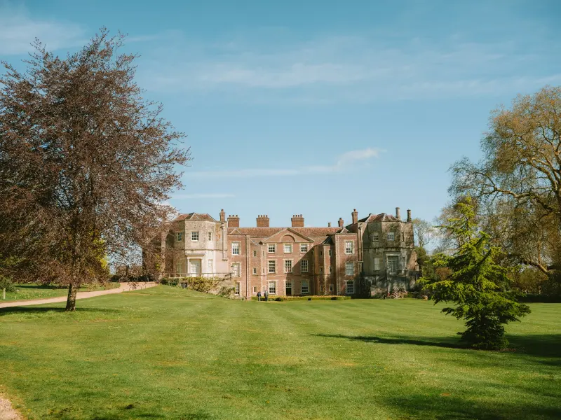 Red-brick manor house across sunlit lawns under a blue summer sky